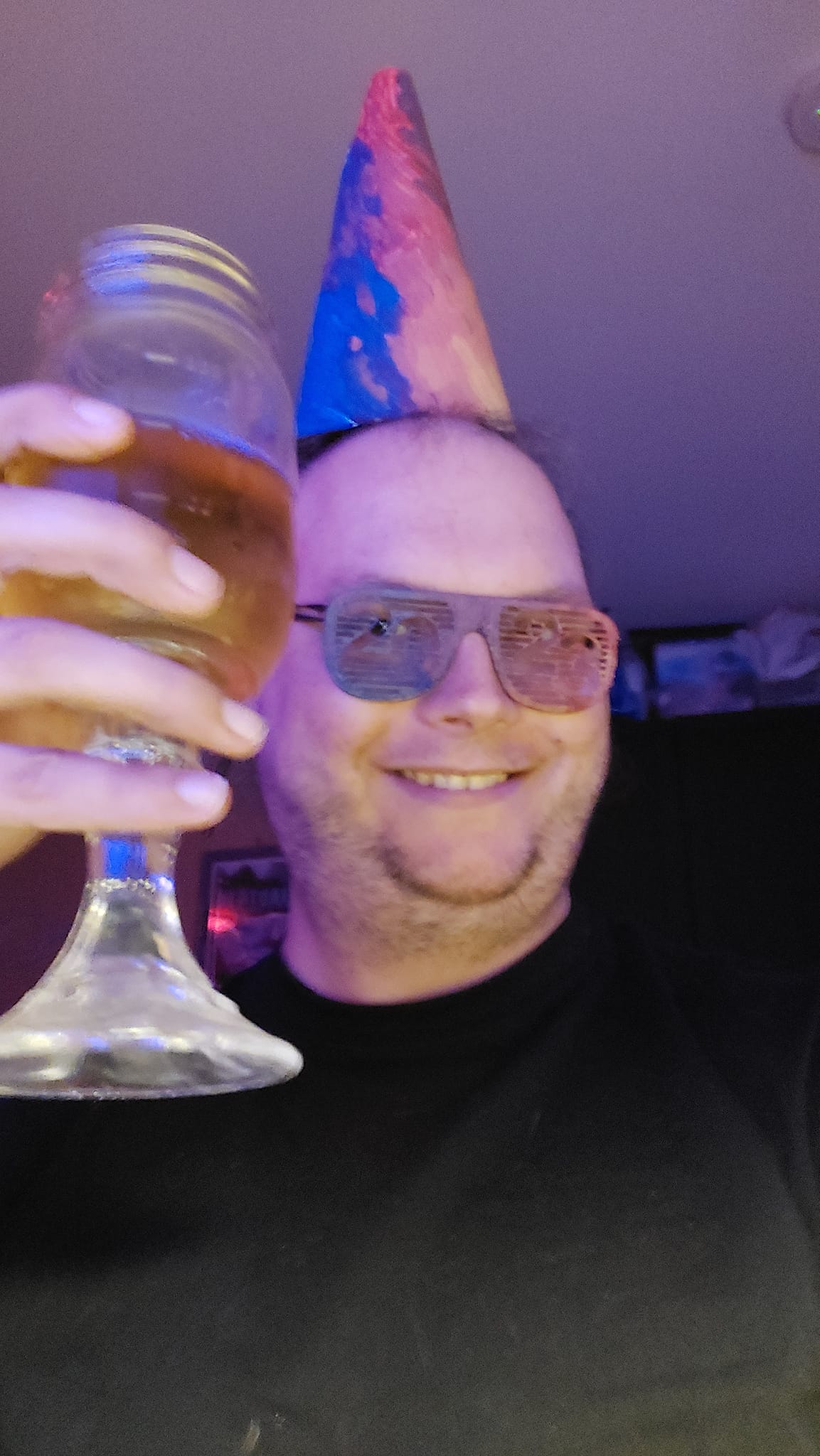 A man sits in a corner, wearing both the hat and painted glasses, he's holding a mason jar wine glass with apple juice in it